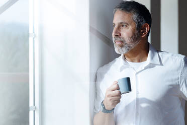 Älterer Geschäftsmann mit Kaffeetasse, der durch das Fenster schaut, während er zu Hause steht - SBOF02553