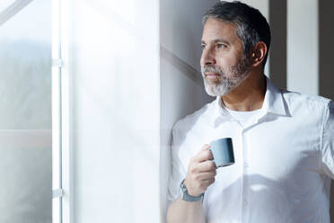 Mature businessman with coffee cup looking through window while standing at home - SBOF02553