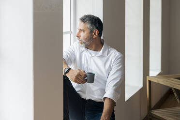 Thoughtful businessman with coffee cup looking through window while sitting at home - SBOF02555
