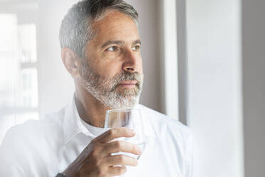 Mature businessman with drinking glass looking away while standing at home - SBOF02557