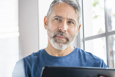 Thoughtful businessman with digital tablet looking away while standing by window at home - SBOF02565