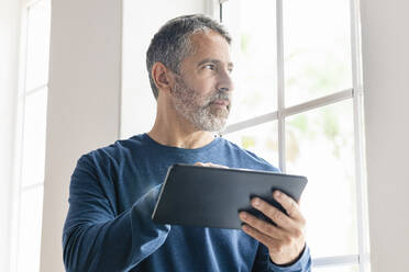 Älterer Geschäftsmann mit digitalem Tablet, der wegschaut, während er zu Hause steht - SBOF02567