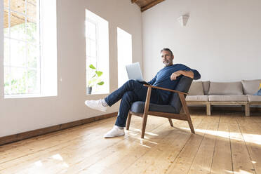 Älterer Geschäftsmann, der einen Laptop benutzt, während er zu Hause auf einem Sessel sitzt - SBOF02574