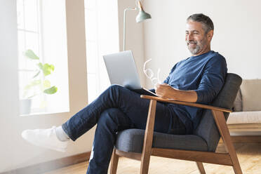 Smiling businessman using laptop while sitting on armchair at home - SBOF02576