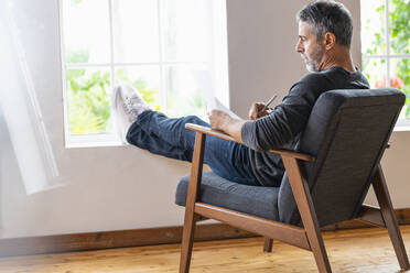 Geschäftsmann schreibt in Papier, während er mit gekreuzten Beinen auf einem Sessel zu Hause sitzt - SBOF02583