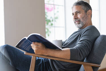 Älterer Geschäftsmann mit Kaffeetasse und Buch, der wegschaut, während er zu Hause sitzt - SBOF02589