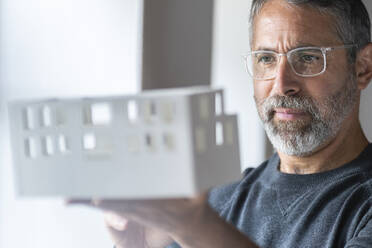 Businessman wearing eyeglasses examining architectural model while standing at home - SBOF02594
