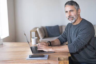 Businessman with laptop looking away while sitting at home office - SBOF02602