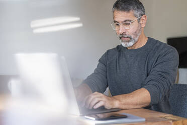 Mature businessman wearing eyeglasses using laptop while working at home - SBOF02604