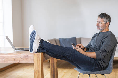 Thoughtful businessman looking away while sitting with mobile phone at home - SBOF02610