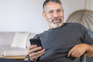 Lächelnder Geschäftsmann mit Brille, der ein Mobiltelefon benutzt, während er zu Hause sitzt - SBOF02611