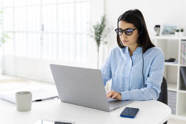 Female customer service representative with headset working on laptop while sitting at office - GIOF10887