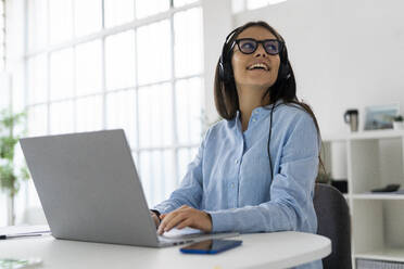 Lächelnde Geschäftsfrau mit Mikrofon-Headset, die wegschaut, während sie mit einem Laptop im Büro sitzt - GIOF10890