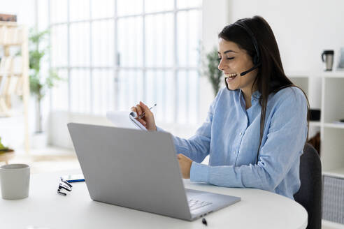 Fröhliche Geschäftsfrau mit Mikrofon-Headset, die in einen Notizblock schreibt, während sie mit einem Laptop im Büro sitzt - GIOF10894