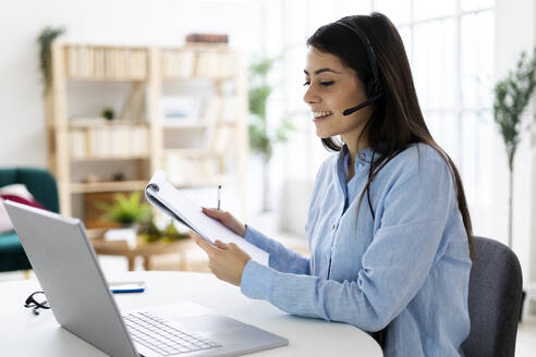 Smiling woman wearing microphone headset holding notepad while working on laptop at office - GIOF10895