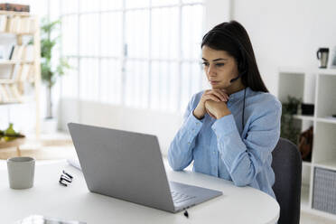 Businesswoman wearing headset using laptop while sitting with hand on chin at office - GIOF10900