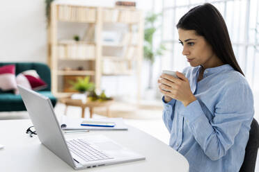 Female entrepreneur drinking coffee while using laptop sitting at office - GIOF10908