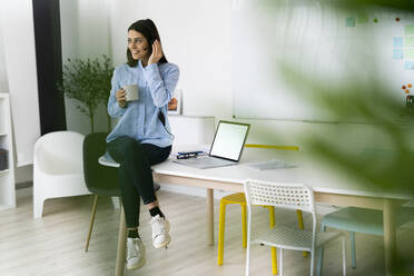 Smiling businesswoman wearing headset drinking coffee while sitting on desk by laptop at office - GIOF10911