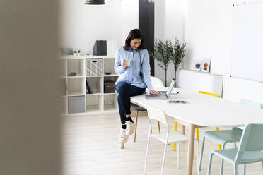 Smiling businesswoman drinking coffee while working on laptop at office - GIOF10914