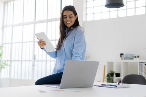 Weibliche Fachkraft hält digitales Tablet, während sie im Büro am Laptop arbeitet - GIOF10915