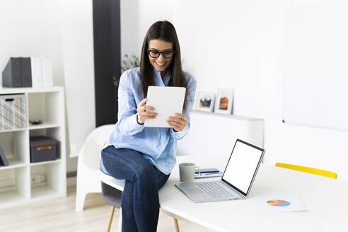 Lächelnde Geschäftsfrau, die an einer Telefonkonferenz auf einem digitalen Tablet teilnimmt, während sie im Büro am Laptop sitzt - GIOF10917