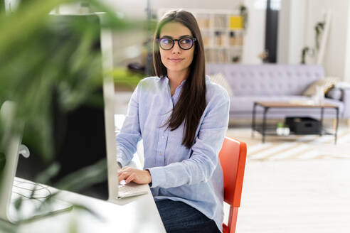 Berufstätige Frau mit Brille, die einen Laptop benutzt, während sie am Schreibtisch im Büro sitzt - GIOF10920