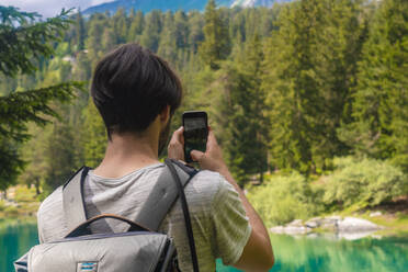 Männlicher Wanderer, der den Caumasee mit seinem Handy fotografiert, während er im Kanton Graubünden steht, Schweiz - TAMF02815