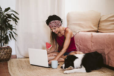 Lächelnde Frau, die einen Laptop benutzt, während sie neben einem Border Collie Hund zu Hause sitzt - EBBF02336