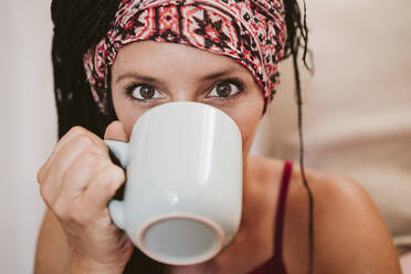 Mittlere erwachsene Frau mit Kopftuch trinkt Kaffee, während sie zu Hause sitzt - EBBF02350