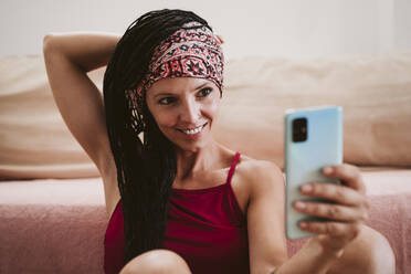 Lächelnde Frau mit Kopftuch, die ein Selfie macht, während sie zu Hause auf dem Sofa sitzt - EBBF02354