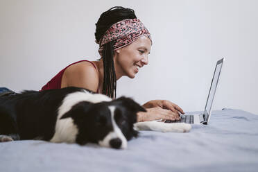 Lächelnde Frau, die einen Laptop benutzt, während sie zu Hause auf dem Bett bei ihrem Haustier liegt - EBBF02359