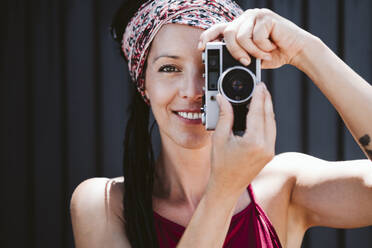 Smiling woman taking photo through camera while standing against wall - EBBF02369