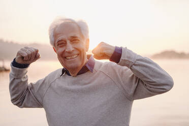 Smiling senior man showing bicep while standing against sky during sunrise - GUSF05027