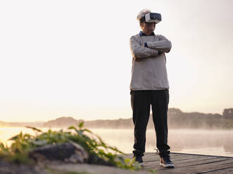 Lächelnder Mann, der ein Virtual-Reality-Headset benutzt, während er bei Sonnenaufgang am Pier steht - GUSF05032