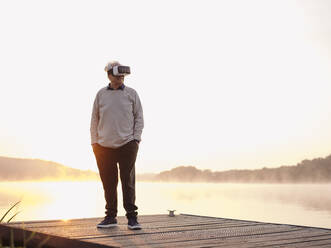 Älterer Mann trägt ein Virtual-Reality-Headset, während er mit den Händen in den Taschen auf einem Pier steht - GUSF05033