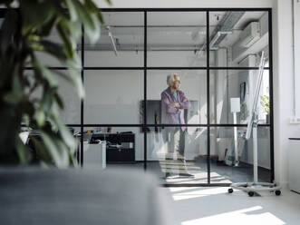 Geschäftsmann mit verschränkten Armen, der wegschaut, während er im Büro steht - GUSF05067