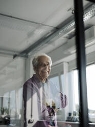 Happy senior entrepreneur standing with arms crossed behind glass wall at office - GUSF05069