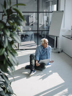 Senior Professional mit Laptop schaut weg, während er im Büro auf dem Boden sitzt - GUSF05077