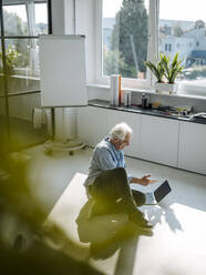 Geschäftsmann im Gespräch mit Videokonferenz über Laptop, während er auf dem Boden im Büro sitzt - GUSF05079