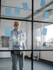 Senior businessman with arms crossed staring while standing by glass wall in office - GUSF05096