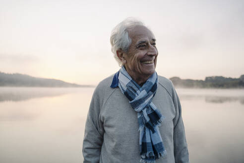 Smiling man wearing scarf looking away while standing against lake - GUSF05119