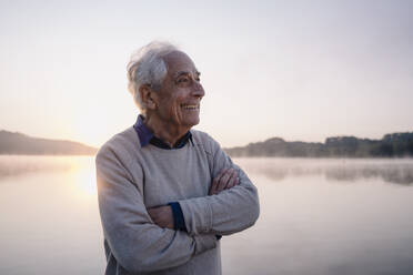 Lächelnder älterer Mann, der wegschaut, während er mit verschränkten Armen am See steht - GUSF05133