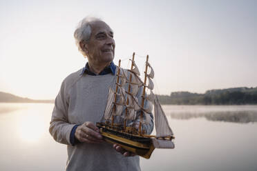 Lächelnder älterer Mann, der ein Spielzeugboot hält, während er am See steht - GUSF05139