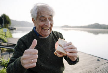 Happy man showing thumbs up while eating sandwich sitting on pier - GUSF05144