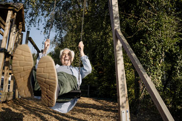 Senior man sticking out tongue while playing on swing at park - GUSF05151