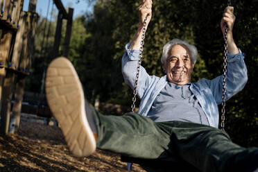 Happy senior man playing while sitting on swing at park - GUSF05152