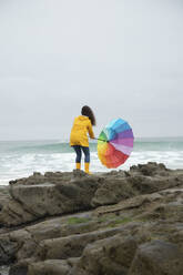 Woman holding umbrella in wind while standing on rock against sea - KBF00699