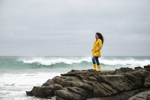 Junge Frau in gelbem Regenmantel, die auf einem Felsen stehend das Meer betrachtet - KBF00700