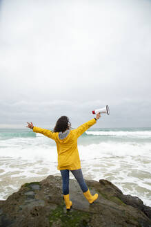 Carefree young woman holding megaphone while standing against sea - KBF00702