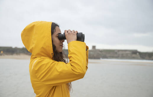 Junge Frau schaut durch ein Fernglas, während sie am Strand steht - KBF00704